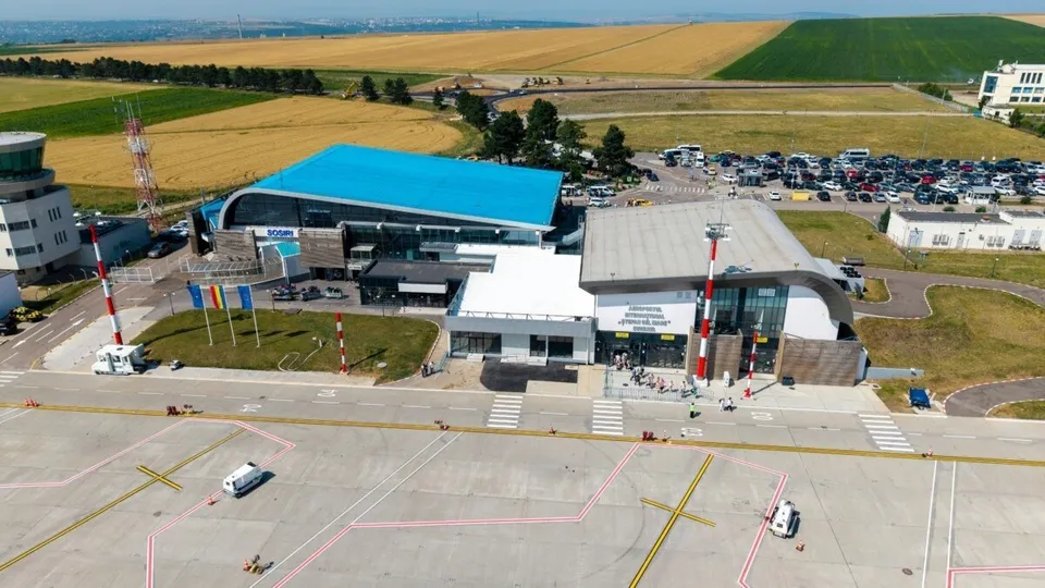 O vedere aeriană a unui terminal de aeroport cu acoperiș albastru, o parcare plină de mașini și o pistă de beton în prim-plan.