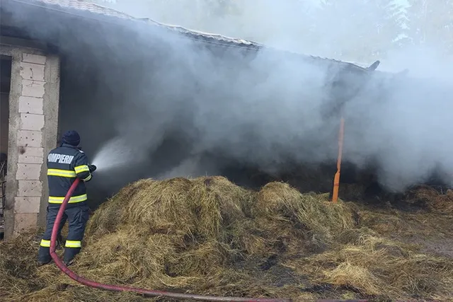 Un pompier în echipament de protecție folosește un furtun pentru a stinge fumul dens de deasupra unui munte de fân lângă o clădire.