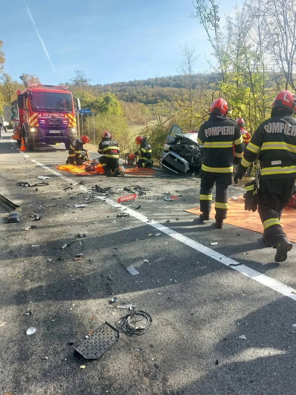 Pompieri în uniforme negre și galbenii stau pe un drum asfaltat lângă un vehicul negru distrus și un camion roșu.