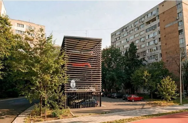 O structură metalică cu lămpi orizontale de parcare este situată între copaci și clădiri de locuințe înalte sub un cer luminos.