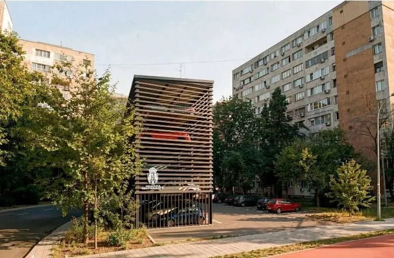 O structură metalică cu lămpi orizontale de parcare este situată între copaci și clădiri de locuințe înalte sub un cer luminos.