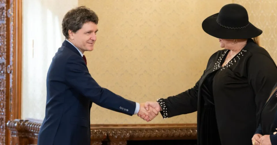 Un bărbat într-un costum închis și o femeie cu pălărie neagră se strâng de mână într-o încăpere cu tapet galben.
