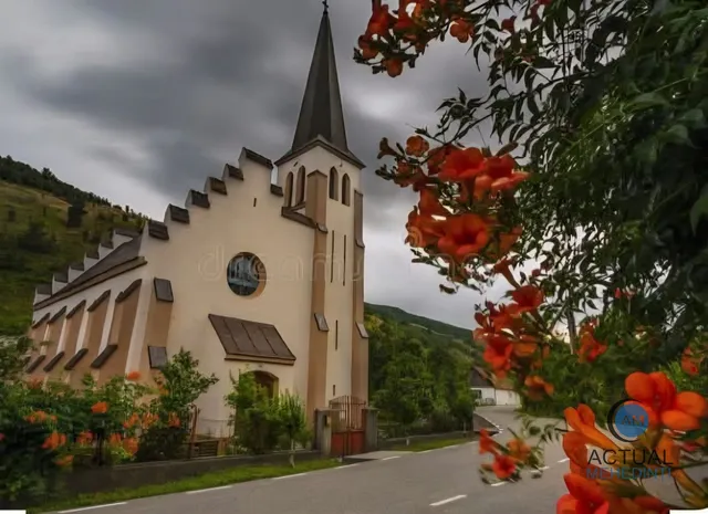 O biserică cu un acoperiș în zigzag și un clopot înalt, încadrată de flori portocalii și un drum asfaltat.