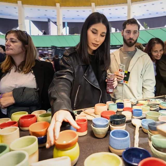 O femeie cu o jachetă de piele neagră întinde mâna spre diverse căni colorate dispuse pe o masă în fața unui grup de persoane.