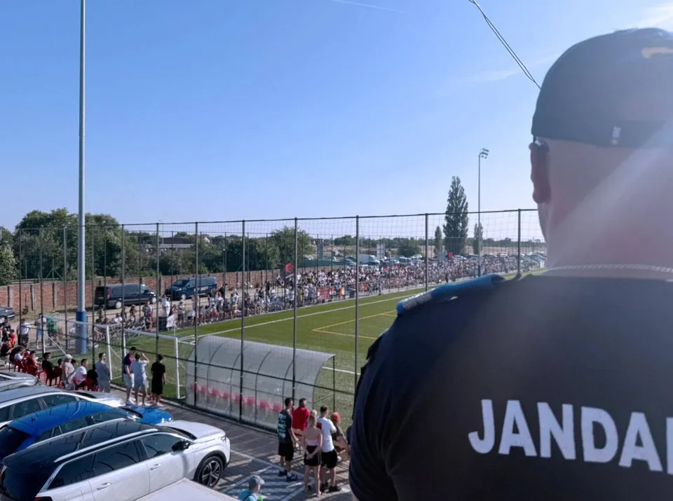 Jandarm în prim-plan privește spre stadionul de fotbal, cu spectatori în tribune și vehicule parcate pe lângă teren.