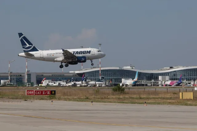 Un avion alb cu însemne albastre zboară deasupra unui terminal de aeroport cu clădiri moderne și alte aeronave parcate în fundal.