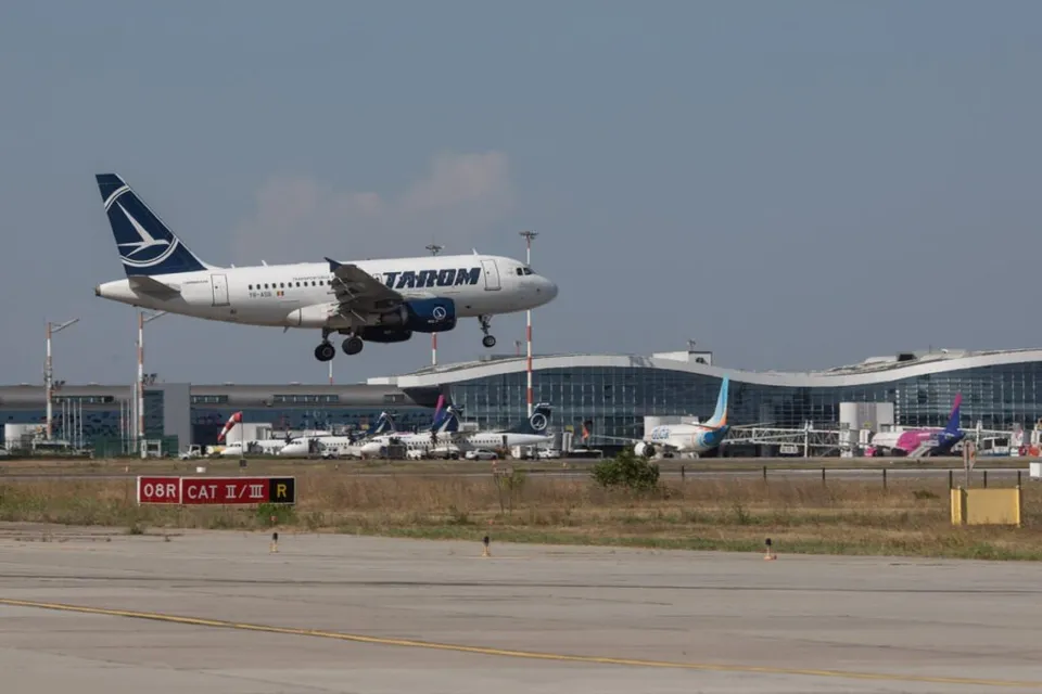 Un avion alb cu însemne albastre zboară deasupra unui terminal de aeroport cu clădiri moderne și alte aeronave parcate în fundal.