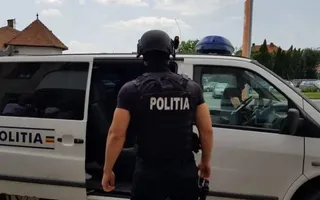 Un agent de poliție cu vestă tactică scrisă pe spate este poziționat lângă o mașină albă cu inscripția politia.