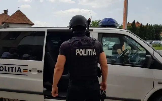 Un polițist cu echipament tactic și cască neagră stă lângă o mașină de poliție albă cu inscripția POLITIA pe spate și pe lateral.