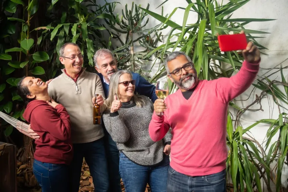 Un grup de cinci adulți zâmbește și face o fotografie de tip selfie în fața unor plante verzi de interior.