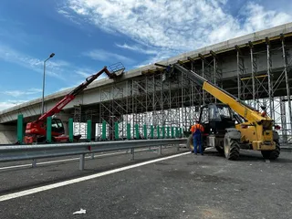 Două utilaje de ridicat cu braț telescopic sunt poziționate pe un drum lângă o structură de beton susținută de schele metalice sub un cer înnorat.