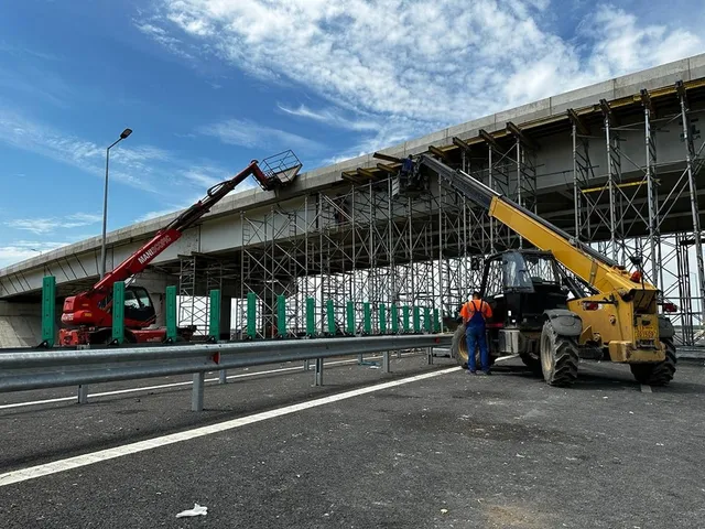 Două utilaje de ridicat cu braț telescopic sunt poziționate pe un drum lângă o structură de beton susținută de schele metalice sub un cer înnorat.