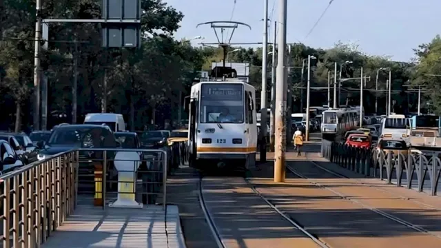 Un tramvai alb cu detalii galbene circulă pe șine într-o zonă urbană, înconjurat de mașini și stâlpi de utilități.