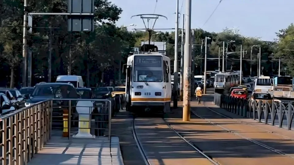 Un tramvai alb cu detalii galbene circulă pe șine într-o zonă urbană, înconjurat de mașini și stâlpi de utilități.