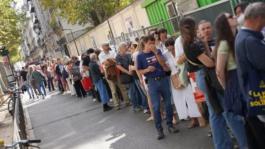 O mulțime de oameni stă la coadă pe un trotuar lângă un gard metalic și un zid de clădiri în lumină naturală.