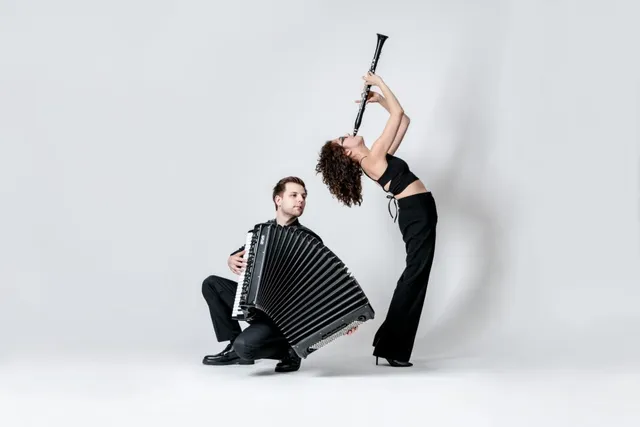 O femeie în haine negre ține un clarinet ridicat, în timp ce un bărbat în haine negre stă ghemuit ținând un acordeon mare.