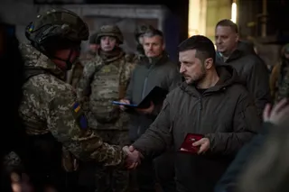 Un bărbat într-o jachetă maro strânge mâna unui soldat în uniformă militară camuflată în fața mai multor persoane.