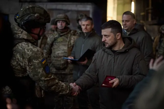 Un bărbat într-o jachetă maro strânge mâna unui soldat în uniformă militară camuflată în fața mai multor persoane.