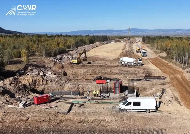 O excavatoare galbenă lucrează pe un teren de pământ lângă un camion mixer și un van alb pe un șantier de construcții.