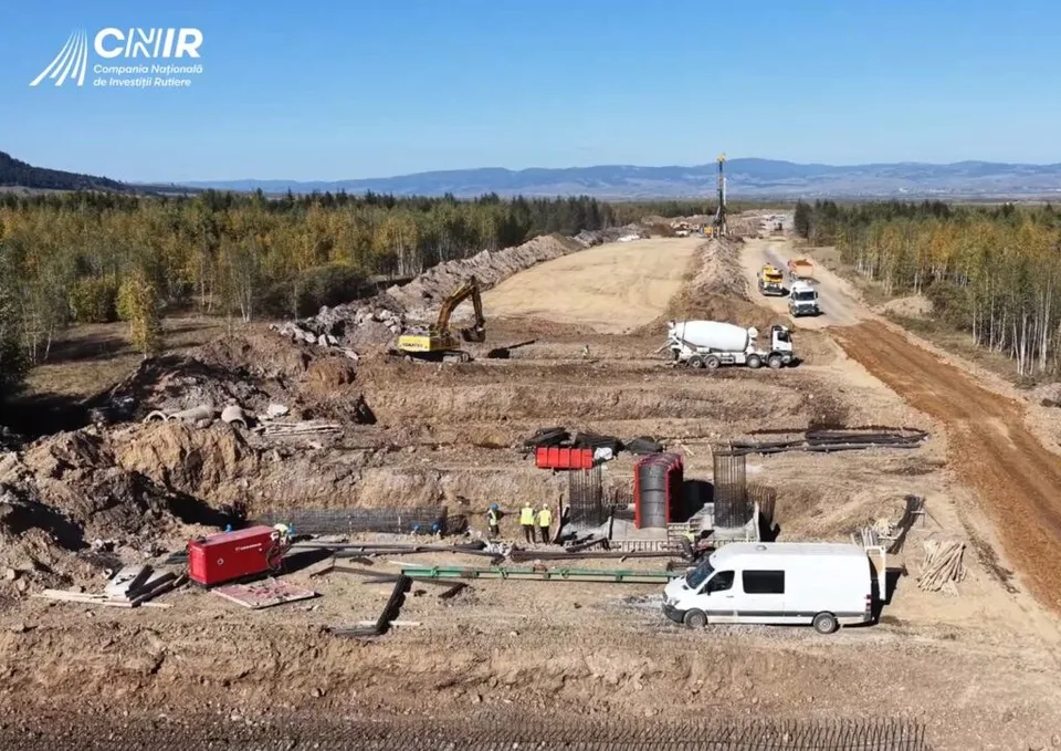O excavatoare galbenă lucrează pe un teren de pământ lângă un camion mixer și un van alb pe un șantier de construcții.