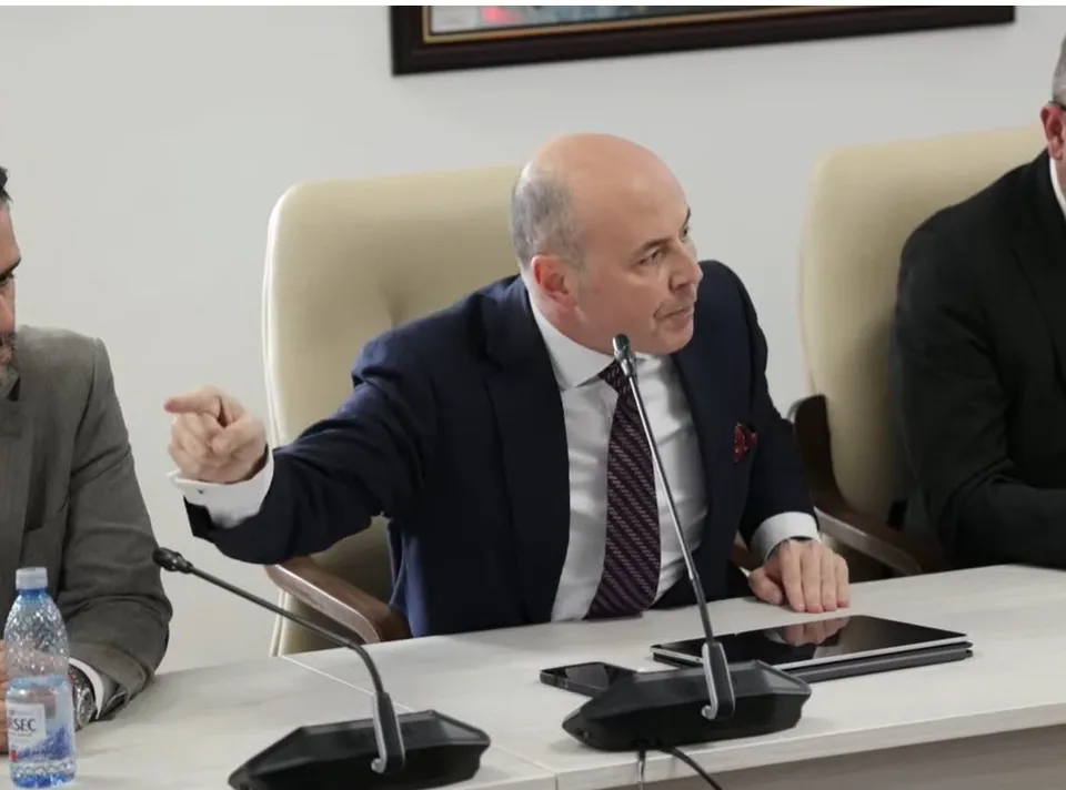 Un bărbat cu pielea deschisă, purtând sacou închis și cravată, stă la o masă albă și arată cu degetul spre stânga.