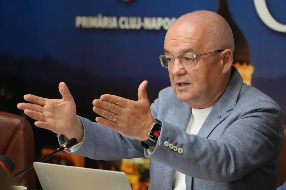 Un bărbat cu ochelari și sacou gri vorbește cu mâinile întinse în față în fața unui laptop.