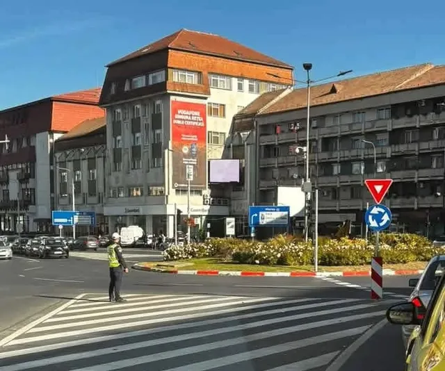 Un om cu vestă reflectorizantă stă pe marginea unui drum lângă un zebru, în fața unor clădiri urbane și a unor semne de circulație.