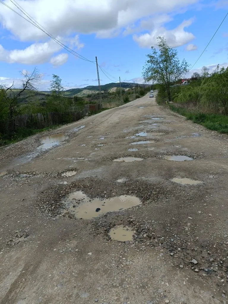 Un drum de țară neasfaltat cu gropi pline de apă se întinde spre dealurile verzi sub un cer cu nori albi.