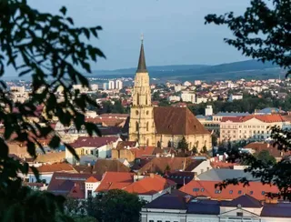 O catedrală cu un turn înalt și ascuțit domină un peisaj urban format din clădiri cu acoperișuri în nuanțe de roșu și portocaliu.