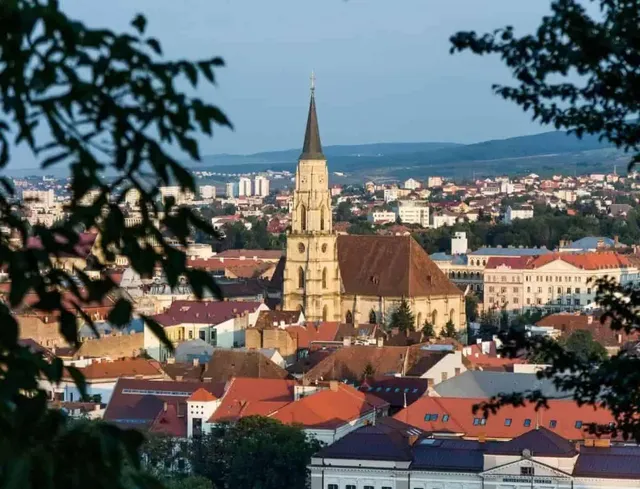 O catedrală cu un turn înalt și ascuțit domină un peisaj urban format din clădiri cu acoperișuri în nuanțe de roșu și portocaliu.