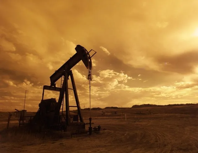 O unitate de pompare de petrol este siluetată pe un fundal de cer cu nori la apusul soarelui într-un peisaj deschis și arid.