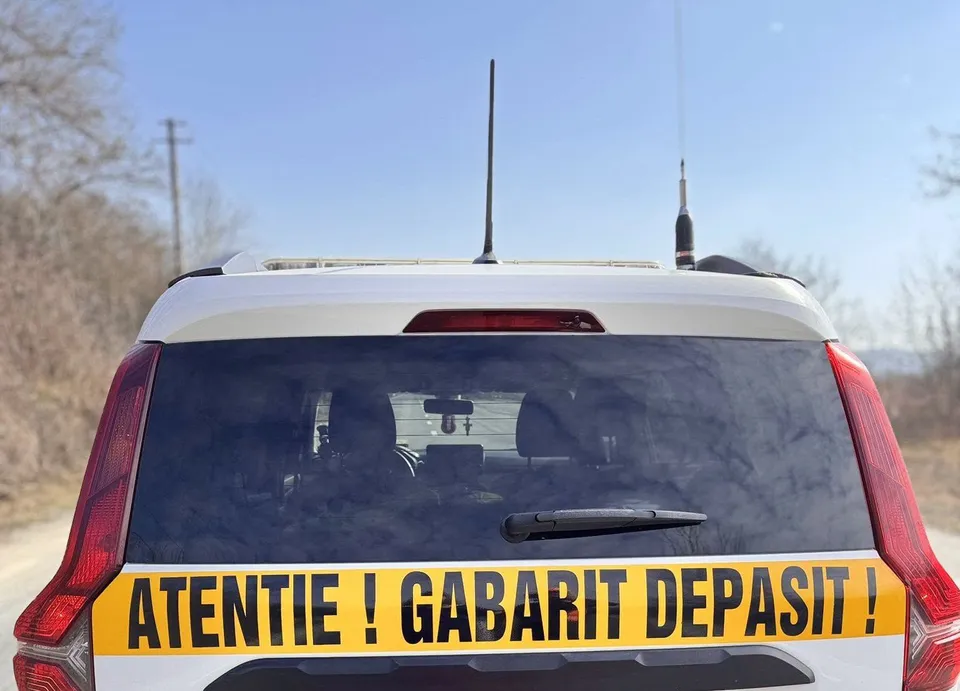 Partea din spate a unui vehicul alb prezintă un autocolant galben pe care scrie text negru cu litere mari.