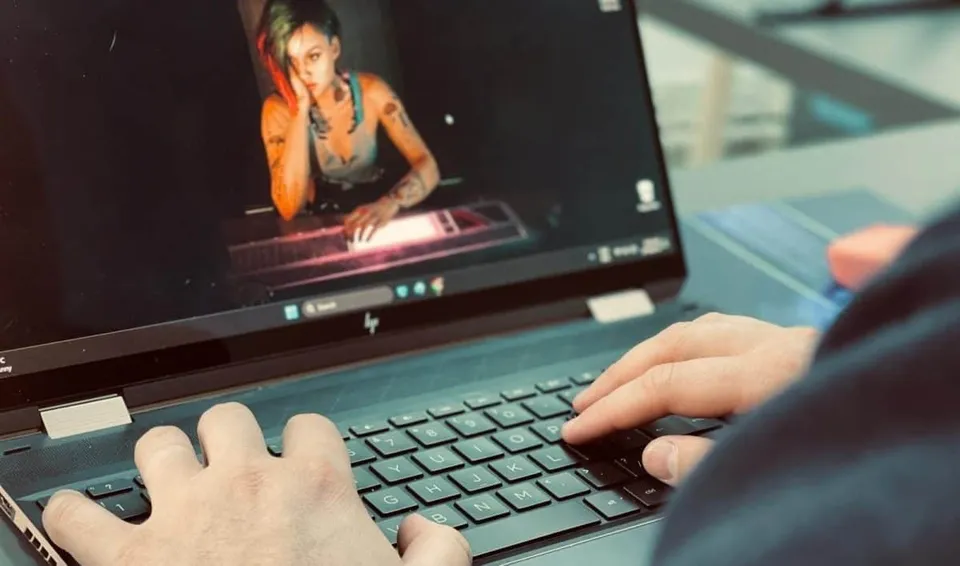 Mâinile unei persoane tastând pe un laptop deschis care afișează o imagine cu o femeie pe ecran.