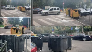 Containere de gunoi de culoare galbenă și neagră sunt amplasate pe un zebru de pe o stradă cu mașini parcate în fundal.