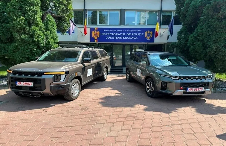 Două vehicule gri sunt parcate pe o curte pavată în fața unei clădiri cu ferestre mari și steaguri colorate.