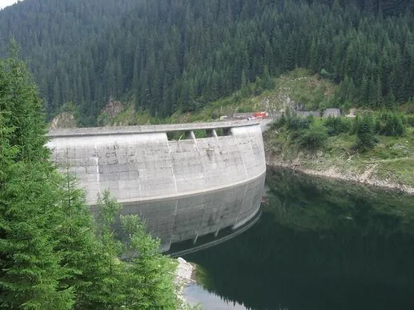 O barajă de beton curbată este situată între un versant împădurit și un lac de apă întunecată.