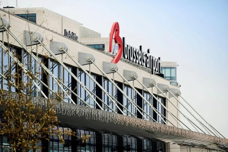 O clădire modernă cu ferestre mari și un logo roșu pe care scrie "brussels airport" sub un cer senin.