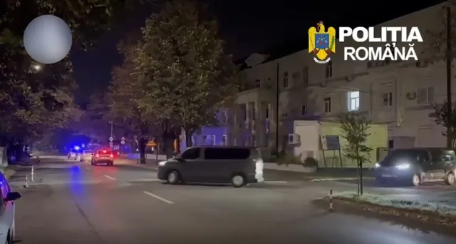Două mașini și un van sunt parcate pe un drum asfaltat lângă clădiri, sub lumini de stradă și un cer de noapte.