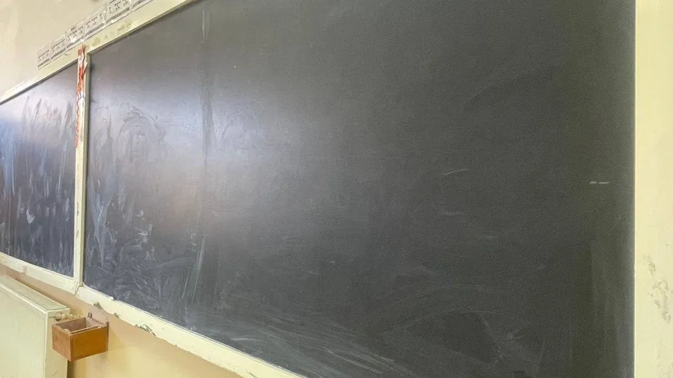 O tablă neagră mare, murdară cu urme de cretă, este montată pe un perete deschis lângă un obiect dreptunghiular maro.