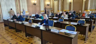 Oamenii stau la mese lungi de conferință în interiorul unei săli cu coloane și podea din lemn, având microfoane și documente în față.