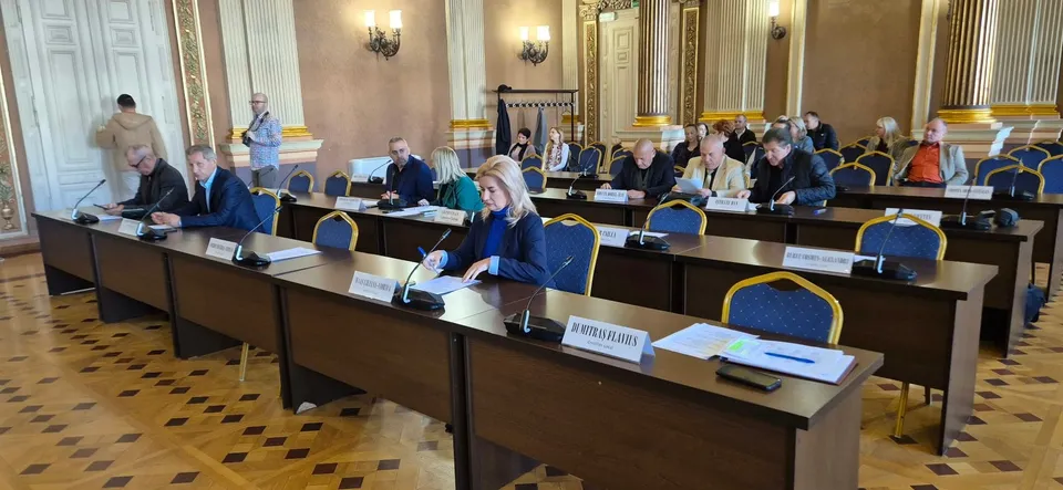 Oamenii stau la mese lungi de conferință în interiorul unei săli cu coloane și podea din lemn, având microfoane și documente în față.