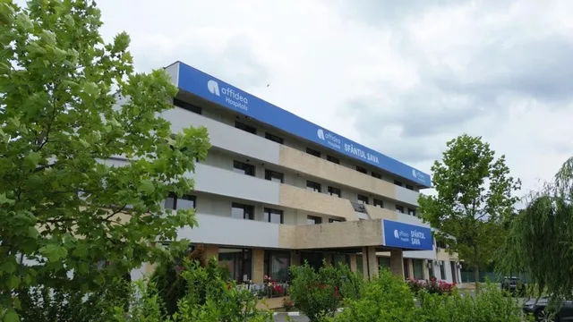O clădire cu mai multe etaje, de culoare albă și gri, prezintă un panou albastru pe partea superioară și un panou albastru deasupra intrării.