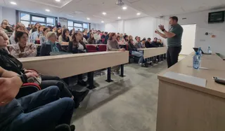 Un bărbat stă în picioare în fața unui grup de oameni așezați la bănci în interiorul unei săli de curs.