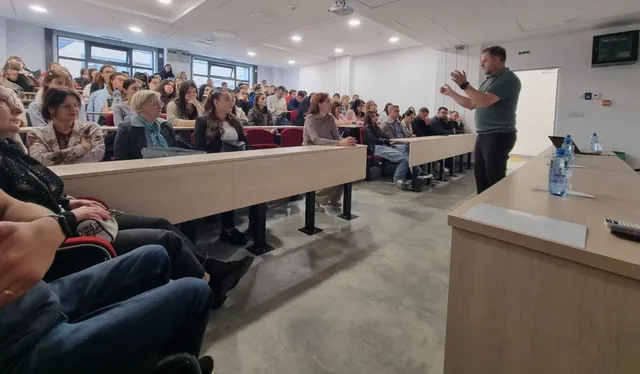 Un bărbat stă în picioare în fața unui grup de oameni așezați la bănci în interiorul unei săli de curs.