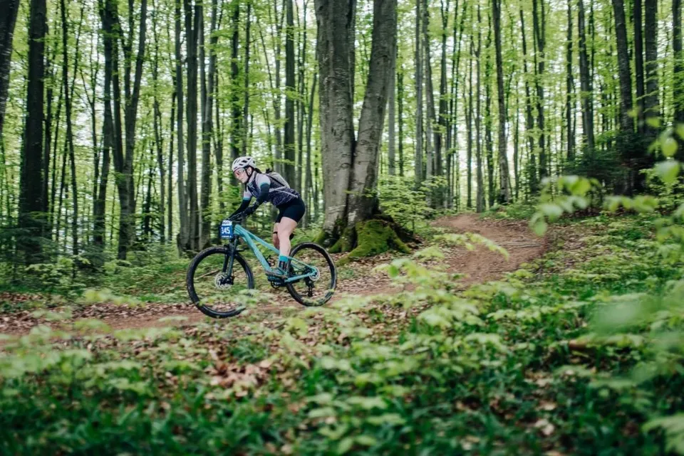 O persoană purtând cască și rucsac pedalează pe un bicicletă montană pe un drum de pământ într-o pădure cu copaci înalți și vegetație verde.