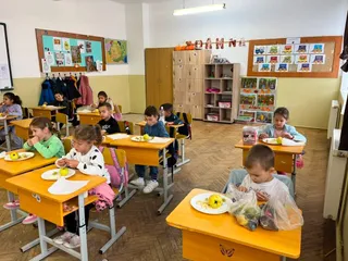 Copii stau la bănci în interiorul unei săli de clasă, având pe mese fructe și obiecte de scris.
