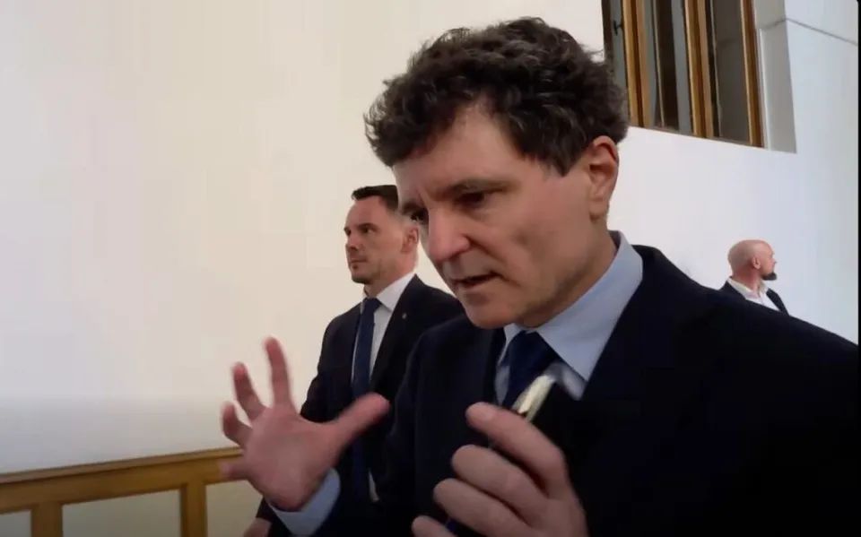Un bărbat cu părul creț și haine închise vorbește cu mâinile ridicate, în timp ce un alt bărbat în costum stă în fundal.