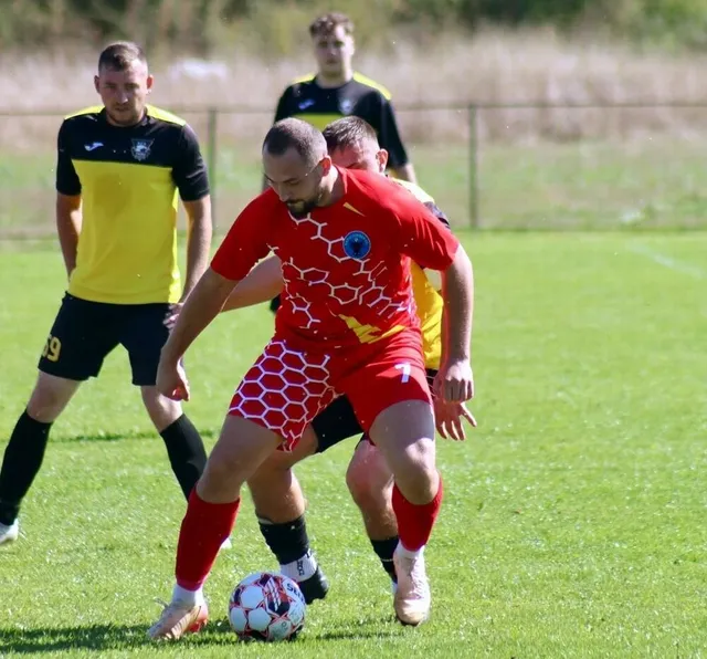 Un jucător de fotbal în echipament roșu cu modele hexagonale conduce mingea pe un teren de iarbă verde în fața altor jucători.