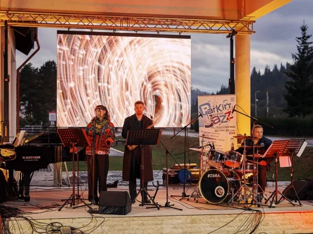 Trei muzicieni execută un concert pe o scenă exterioară în fața unui ecran mare care afișează un model circular de lumină.