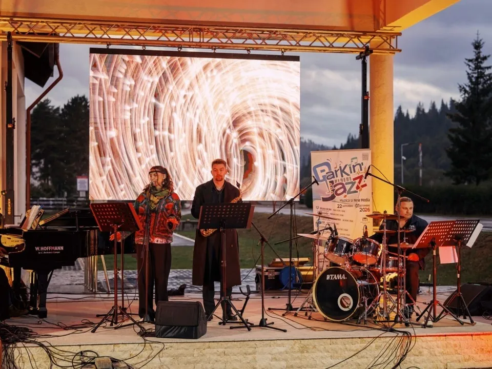 Trei muzicieni execută un concert pe o scenă exterioară în fața unui ecran mare care afișează un model circular de lumină.